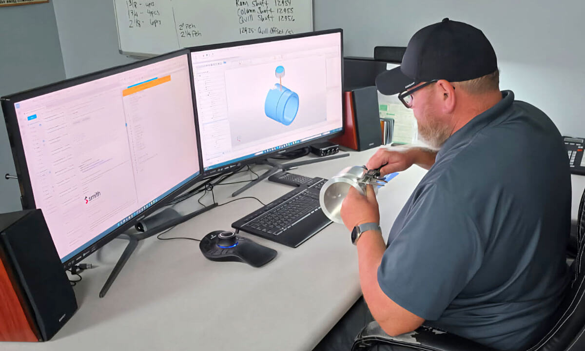 Man sitting in front of two computer screens with a part in his hands. He is creating a computer model of the part.
